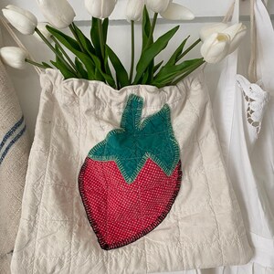 May include: A cream-colored quilted fabric bag with a large strawberry applique. The strawberry has a red dotted body and a green leaf top. The bag is filled with white tulips.