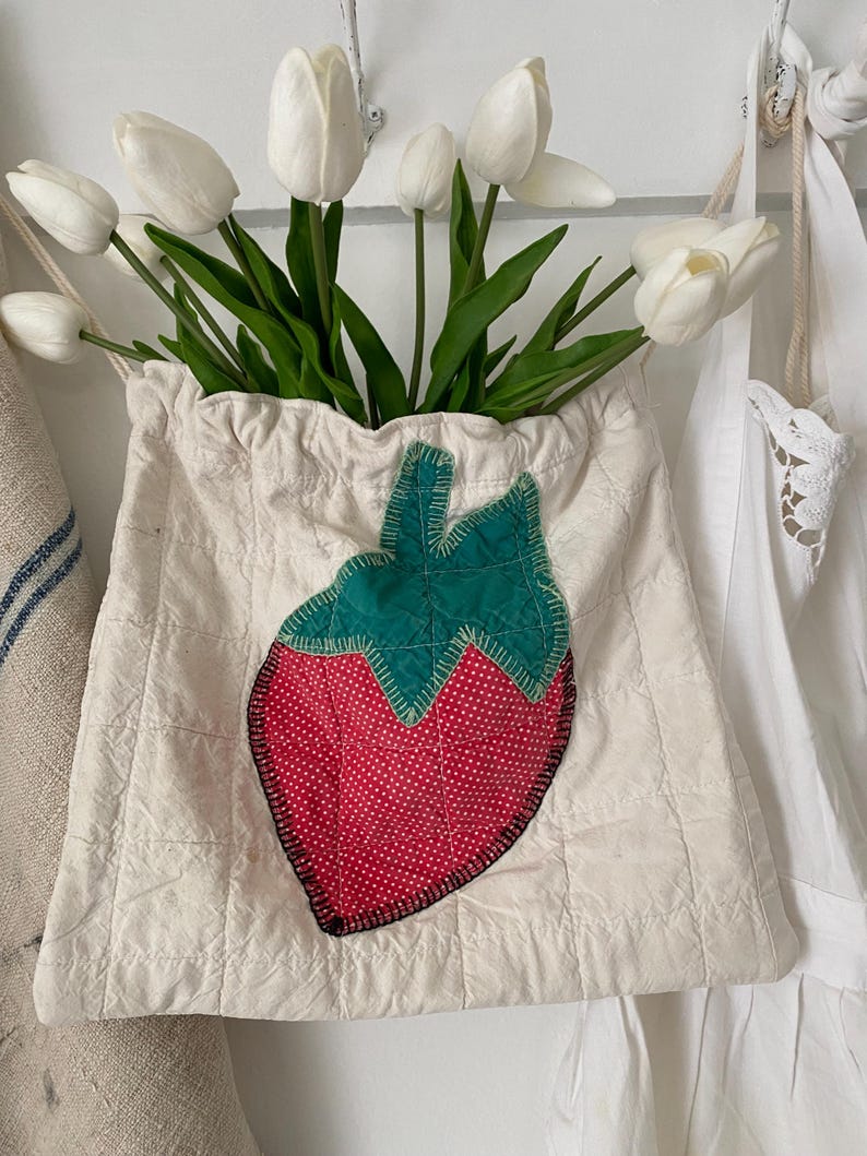 May include: A cream-colored quilted fabric bag with a large strawberry applique. The strawberry has a red dotted body and a green leaf top. The bag is filled with white tulips.