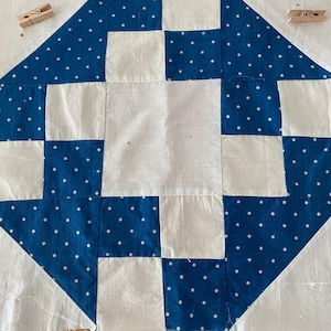 May include: A white quilt block with a blue and white polka dot pattern. The block is made up of nine squares, with the center square being white and the surrounding squares being blue with white polka dots.