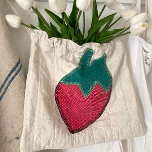 May include: A cream-colored fabric bag with a quilted design and a large strawberry applique. The strawberry is red with white polka dots and a green top. The bag is filled with white tulips.