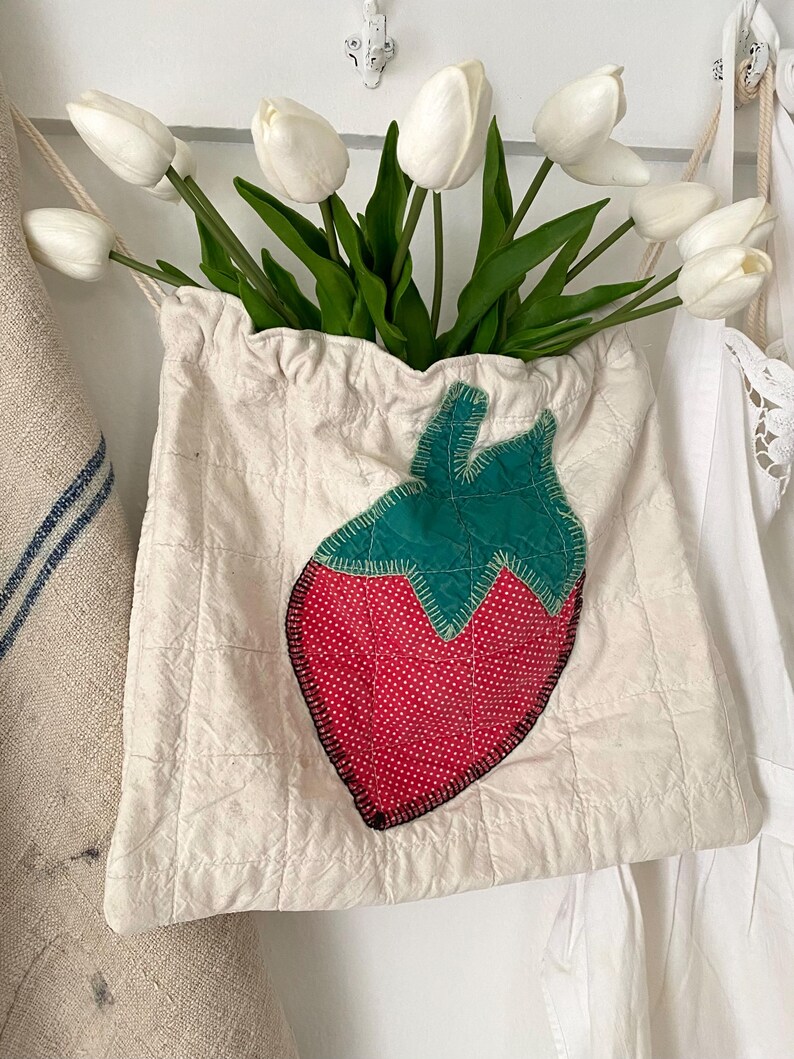 May include: A cream-colored fabric bag with a quilted design and a large strawberry applique. The strawberry is red with white polka dots and a green top. The bag is filled with white tulips.