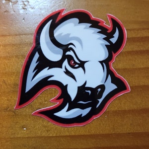 May include: A sticker featuring a stylized buffalo head. The buffalo is white with black and gray accents, a red eye, and a red outline. The sticker is on a wooden surface.