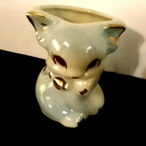 May include: A small, light blue ceramic planter shaped like a cat with gold accents. The cat is sitting with its head down and its paws tucked in.