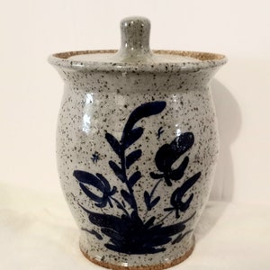 May include: A white ceramic jar with a lid, decorated with a blue floral design. The jar has a speckled glaze and a brown rim.