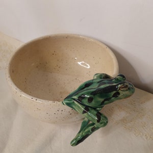 May include: A white ceramic bowl with green and black speckled glaze. A green and black frog figurine is perched on the rim of the bowl.