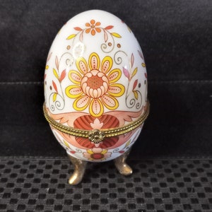 以下が含まれることがあります： オレンジ、イエロー、ピンクの色の花柄が施された、白い磁器製の卵型の小物入れ。金色の縁取りと3本の金色の足がついている。