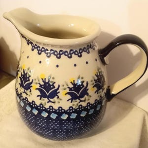 Peut inclure: Un pichet en céramique avec une base de couleur crème et une anse bleu foncé. Le corps est orné d'un motif répétitif de fleurs stylisées bleu foncé, de points jaunes et de feuilles vertes, avec une base bleu foncé à pois blancs.