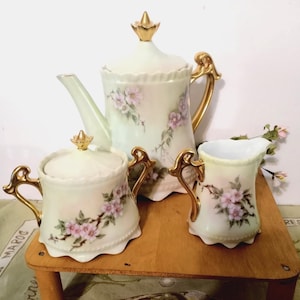 May include: A vintage tea set comprising a teapot, sugar bowl, and creamer. The cream-coloured set features gold accents and delicate pink floral patterns. The teapot has a lid with a gold finial, and the handles are gold.