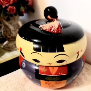 Puede incluir: Una muñeca rusa tradicional, o 'kokeshi', con una cara pintada de negro y amarillo y un kimono rojo y azul. La muñeca está decorada con motivos florales y tiene una tapa negra con un borla roja y blanca.