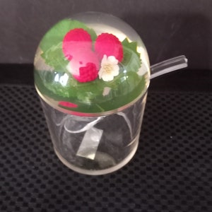 Peut inclure: Un distributeur de sucre en verre transparent avec un couvercle en dôme. Le dôme présente trois framboises en plastique rouge et une fleur en plastique blanche. Le distributeur est doté d'une cuillère en plastique transparente.