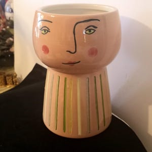 Pode incluir: Um vaso de cerâmica rosa com um design caprichoso. O vaso apresenta um rosto com contornos pretos, olhos verdes e bochechas vermelhas. A base tem listras verticais verdes e brancas. Um item decorativo único.