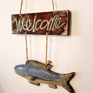 May include: A ceramic welcome sign with a brown wooden background and white lettering. The sign is hanging from a string and has a blue and brown ceramic fish hanging below it.
