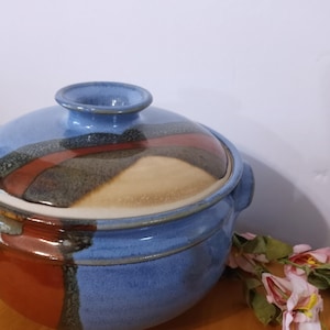 Puede incluir: Una cacerola de cerámica azul y marrón con tapa. La cacerola tiene un diseño abstracto único con vetas marrones, rojas y blancas.