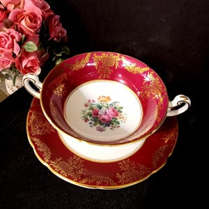 May include: A vintage soup bowl and saucer set. The bowl and saucer are a deep red with gold filigree designs. The interior of the bowl features a floral bouquet. The set has white accents and curved handles.