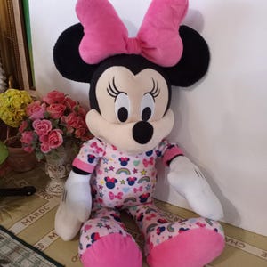 K&ouml;nnte beinhalten: Ein Pl&uuml;schtier von Minnie Mouse, das einen rosa Schlafanzug mit Regenbogen- und Sternenmuster tr&auml;gt. Sie hat eine rosa Schleife auf dem Kopf und schwarze Ohren.