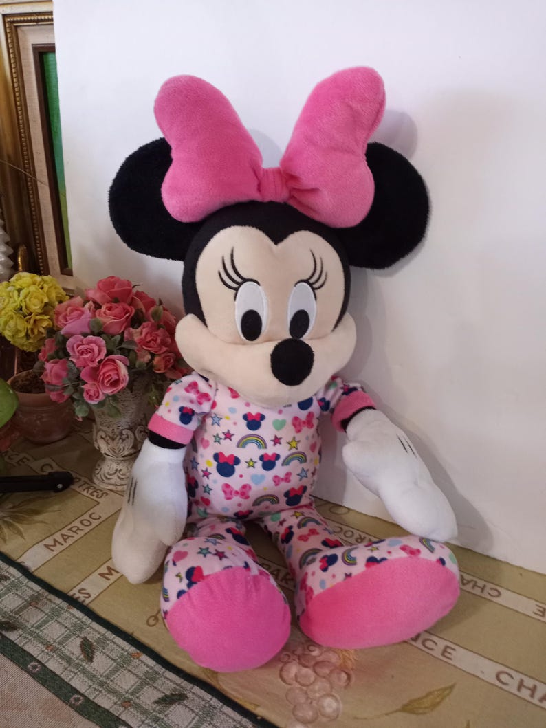 K&ouml;nnte beinhalten: Ein Pl&uuml;schtier von Minnie Mouse, das einen rosa Schlafanzug mit Regenbogen- und Sternenmuster tr&auml;gt. Sie hat eine rosa Schleife auf dem Kopf und schwarze Ohren.