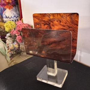 May include: A clear acrylic desk organizer with a rectangular base and a vertical support holding a brown and orange marbled acrylic panel. The organizer has a second panel in front. The design is a decorative office accessory.