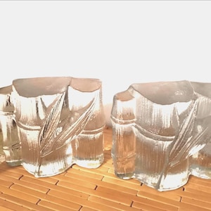 Puede incluir: Dos candelabros de vidrio transparente con diseño de bambú. Los candelabros tienen forma de tallos de bambú con detalles de hojas grabadas. Se encuentran sobre una esterilla de bambú marrón claro.