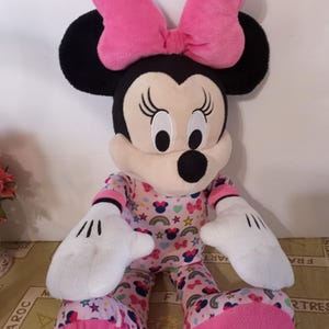 K&ouml;nnte beinhalten: Ein gro&szlig;es Pl&uuml;schtier von Minnie Mouse, das einen rosa Schlafanzug mit Regenbogen, Sternen und Minnie Mouse-Druck tr&auml;gt. Sie hat eine rosa Schleife auf dem Kopf und wei&szlig;e Handschuhe.