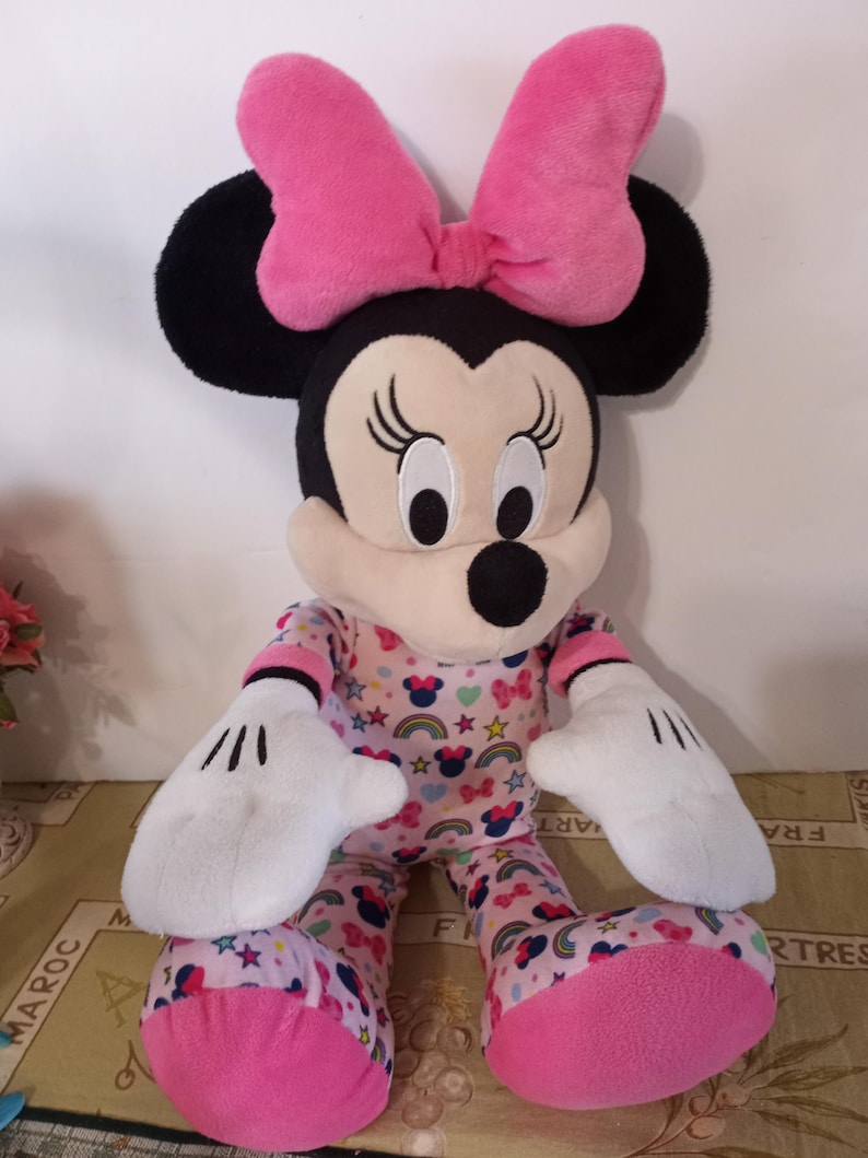K&ouml;nnte beinhalten: Ein gro&szlig;es Pl&uuml;schtier von Minnie Mouse, das einen rosa Schlafanzug mit Regenbogen, Sternen und Minnie Mouse-Druck tr&auml;gt. Sie hat eine rosa Schleife auf dem Kopf und wei&szlig;e Handschuhe.