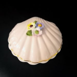 May include: A pale pink ceramic trinket box shaped like a seashell with a scalloped surface. The lid features a cluster of small, sculpted flowers in yellow, blue, and white with green leaves. The edge of the lid is trimmed with a thin gold line.