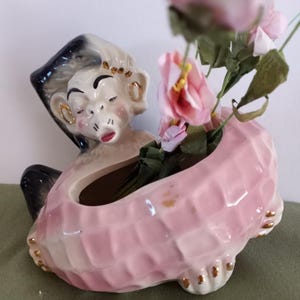 May include: A pink ceramic planter shaped like a peanut with a monkey figurine on top. The monkey has black hair, gold earrings, and a closed eye. The planter has gold accents and is filled with pink flowers.