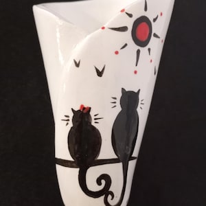 May include: White ceramic vase featuring a black cat and kitten design. The vase has a red sun with black rays and red dots. The cats are perched on a branch with their tails curled.