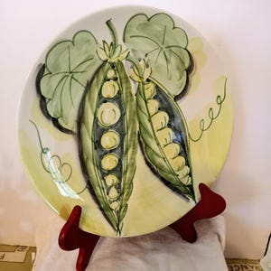 May include: A decorative ceramic plate featuring a hand-painted design of two open pea pods with visible peas. The pods are green with yellow peas, set against a pale yellow and white background. The plate is displayed on a red stand.