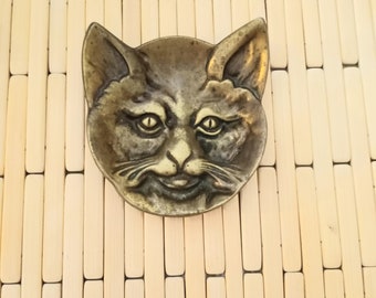 May include: A brass cat head trinket with a detailed face and whiskers. The cat has a mischievous grin and is made of a dark, aged brass.