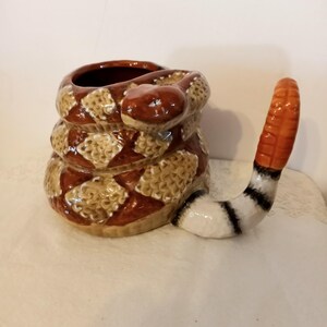 Puede incluir: Una taza de cerámica con forma de serpiente enroscada, con un patrón de diamantes marrón y beige. La cabeza de la serpiente forma la parte superior de la taza, y la cola es el asa. El asa es blanca con rayas negras y una punta naranja.
