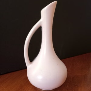 May include: A small, pale pink ceramic vase with a slender neck and a rounded body. The vase has a single handle.