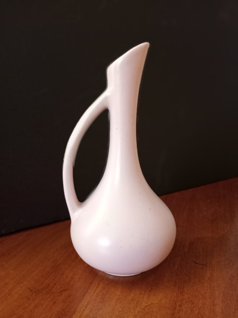 May include: A small, pale pink ceramic vase with a slender neck and a rounded body. The vase has a single handle.