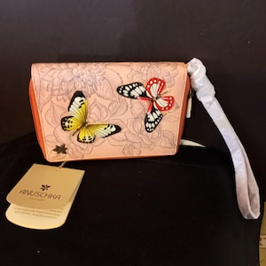 Puede incluir: Un bolso de mano rosa claro con un diseño floral y detalles de mariposas tridimensionales en amarillo, rojo y negro. El bolso tiene un borde naranja y una correa de muñeca blanca. Una etiqueta con la marca "ANUSCHKA" es visible.
