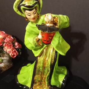 May include: A ceramic figurine of a woman in a green and gold dress and hat, holding a red and black lantern. The figurine has a detailed face and is posed in a graceful stance. The base is black.