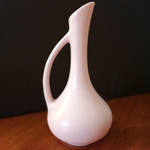 May include: A small, pale pink ceramic vase with a slender neck and a curved handle.