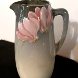 Puede incluir: Jarra de cerámica con la parte superior y el asa de color gris verdoso, que se desvanece hasta una base blanco crema. Dos diseños florales rosas en la parte delantera. La jarra tiene un pico.