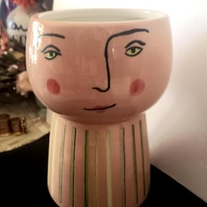 May include: A pink ceramic planter with a painted face design. The planter features a rounded top with a face and a striped base. The face has black outlines, green eyes, and red cheeks. The base has vertical stripes of green and white.