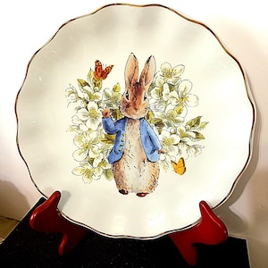 May include: A white ceramic plate with a scalloped edge and gold trim. The plate features a watercolor illustration of a rabbit in a blue jacket surrounded by white flowers and butterflies. The plate is displayed on a red stand.
