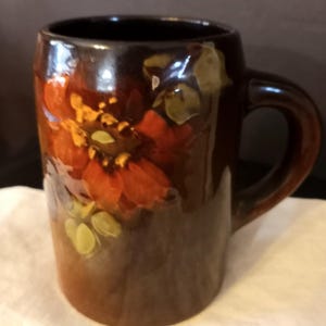May include: A brown ceramic mug with a floral design. The mug features a dark brown exterior with a glossy finish and a large handle. The floral design includes orange, red, yellow, and green colors. The mug is approximately 4 inches tall.
