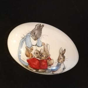 Puede incluir: Un huevo decorativo ovalado blanco con una ilustración en acuarela de conejos. Los conejos están representados en varias poses, algunos con chaquetas rojas y otros con ropa azul. La obra de arte recuerda a un libro infantil.