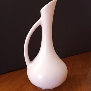 May include: A pale pink ceramic vase with a slender neck and a rounded body. The vase has a single handle and a slightly flared opening.