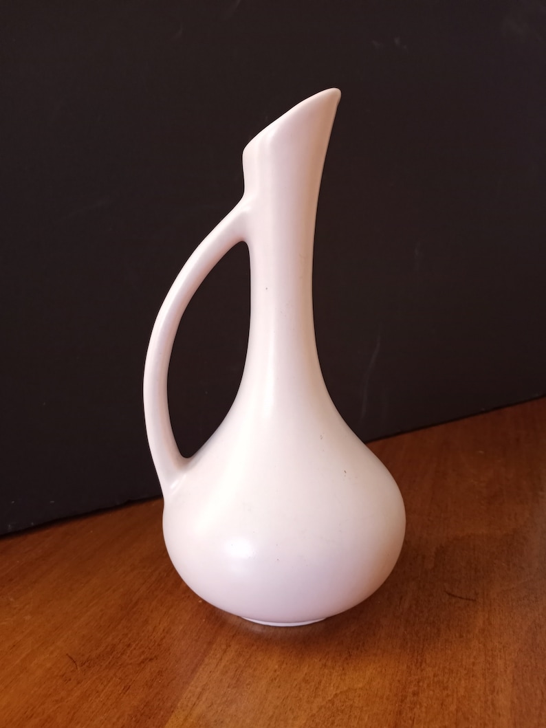 May include: A pale pink ceramic vase with a slender neck and a rounded body. The vase has a single handle and a slightly flared opening.