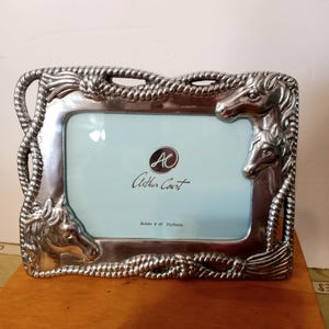 May include: Silver-toned metal picture frame with a rectangular opening. The frame features horse head details at the corners and along the sides, with a rope-like border. The frame is marked with the text "Arthur Court" and "Holds 4 x 6 Picture."
