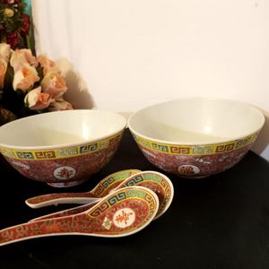 May include: Two ceramic bowls and two matching spoons featuring a red background with intricate gold and white patterns. The rims are decorated with a yellow and blue geometric border. Each piece displays a circular medallion with Chinese characters.