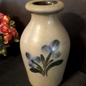 Puede incluir: Un jarrón de gres beige con borde redondeado y cuello estrecho. El cuerpo del jarrón presenta un diseño floral pintado a mano que consiste en dos flores de color azul claro con hojas y tallos verdes centrados en la parte frontal del objeto.