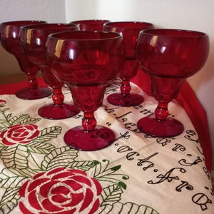 May include: Set of six red glass goblets with a textured design. The goblets are on a white tablecloth with red embroidered roses.