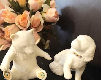 May include: Two white porcelain animal figurines. One is a seated bear with gold and green accents on its paws. The other is a standing dog with a gold and blue collar. Both figures have a smooth, glossy finish and are positioned side by side.
