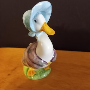 May include: A ceramic duck figurine wearing a pale blue bonnet and a purple and grey vest. The duck has a white body and a yellow beak, standing on a green base with yellow feet. The figurine is a decorative item.