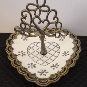 May include: A gold-tone metal ring holder with a heart-shaped base and a decorative tree-shaped top. The base is white with a heart design and floral accents.