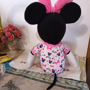 K&ouml;nnte beinhalten: Ein Pl&uuml;schtier von Minnie Mouse, das einen rosa Schlafanzug mit einem Regenbogen-, Sterne- und Herzmuster tr&auml;gt. Sie hat eine rosa Schleife auf dem Kopf und schwarze Ohren.